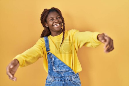Téléchargez les photos : Femme africaine debout sur fond jaune regardant la caméra souriant à bras ouverts pour étreindre. expression joyeuse embrassant le bonheur. - en image libre de droit