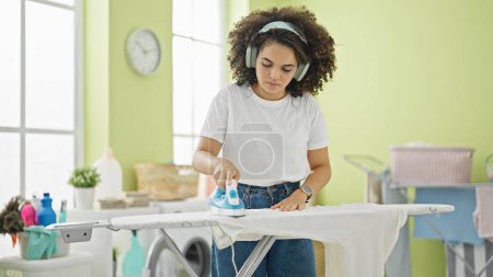 Foto de Joven hermosa mujer hispana escuchando música planchando ropa en la lavandería - Imagen libre de derechos
