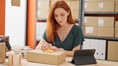 Photo for Young redhead woman ecommerce business worker writing on reminder paper using touchpad at office - Royalty Free Image