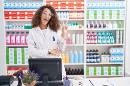 Téléchargez les photos : Femme hispanique avec les cheveux bouclés travaillant à la pharmacie pharmacie souriant avec le visage heureux clin d'oeil à la caméra faisant signe de victoire avec les doigts. numéro deux. - en image libre de droit