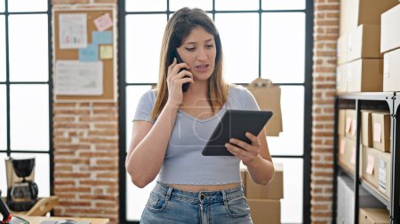 Foto de Mujer rubia joven comercio electrónico trabajador de negocios utilizando touchpad hablando en el teléfono inteligente en la oficina - Imagen libre de derechos