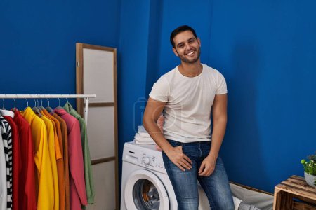 Téléchargez les photos : Jeune homme hispanique souriant confiant assis sur la machine à laver à la buanderie - en image libre de droit