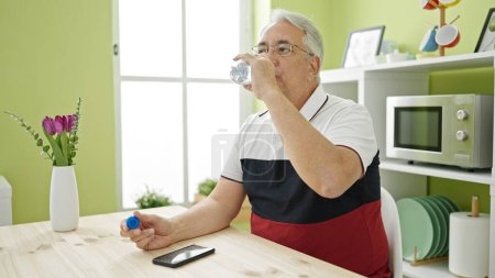 Téléchargez les photos : Homme d'âge moyen avec les cheveux gris assis sur la table boire de l'eau à la salle à manger - en image libre de droit