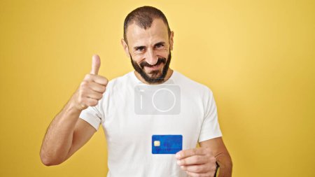 Téléchargez les photos : Jeune homme hispanique tenant la carte de crédit avec le pouce vers le haut geste sur fond jaune isolé - en image libre de droit