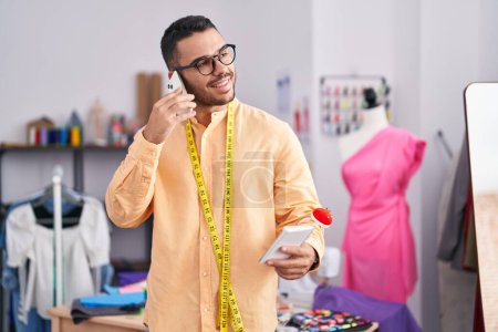 Téléchargez les photos : Jeune homme hispanique tailleur parler sur le portable de lecture smartphone au magasin de tailleur - en image libre de droit