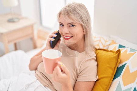 Photo for Young blonde woman talking on smartphone drinking coffee at bedroom - Royalty Free Image