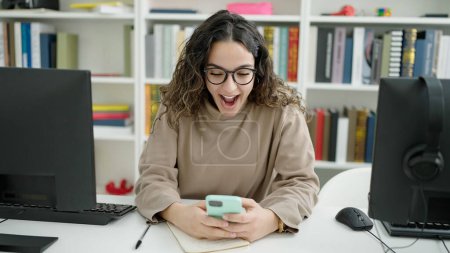 Téléchargez les photos : Jeune belle étudiante hispanique utilisant un smartphone avec expression surprise à la bibliothèque de l'université - en image libre de droit