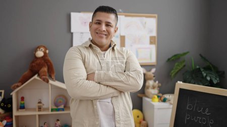 Photo for Young hispanic man preschool teacher smiling confident standing with arms crossed gesture at kindergarten - Royalty Free Image