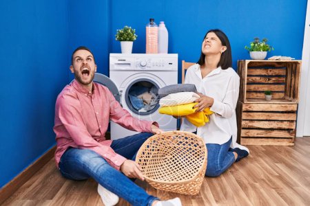 Photo for Young couple doing laundry at home angry and mad screaming frustrated and furious, shouting with anger looking up. - Royalty Free Image