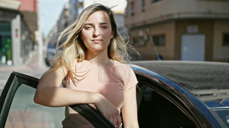 Téléchargez les photos : Jeune femme blonde penchée sur la porte de la voiture avec une expression détendue dans la rue - en image libre de droit