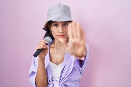 Téléchargez les photos : Jeune femme hispanique chantant chanson à l'aide d'un microphone sur fond rose avec la main ouverte faisant stop signe avec expression sérieuse et confiante, geste de défense - en image libre de droit