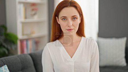 Téléchargez les photos : Jeune femme rousse assise sur un canapé avec un visage sérieux à la maison - en image libre de droit