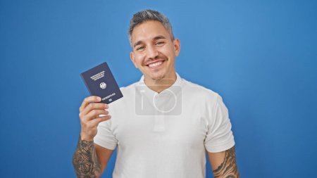 Téléchargez les photos : Jeune homme hispanique souriant confiant tenant passeport deutschland sur fond bleu isolé - en image libre de droit