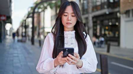 Téléchargez les photos : Jeune femme chinoise utilisant un smartphone avec une expression sérieuse à la rue - en image libre de droit