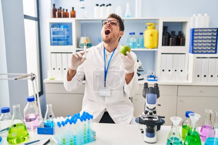 Photo for Young hispanic man working at scientist laboratory holding apple angry and mad screaming frustrated and furious, shouting with anger looking up. - Royalty Free Image