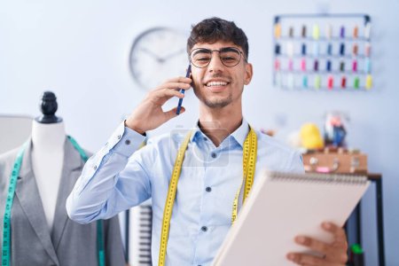 Téléchargez les photos : Jeune homme hispanique tailleur parler sur smartphone regarder carnet à tailleur boutique - en image libre de droit