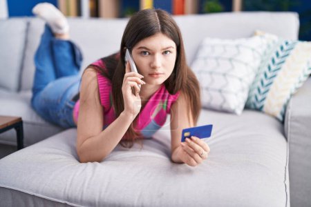Photo for Young caucasian woman talking on smartphone holding credit card lying on sofa at home - Royalty Free Image