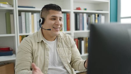 Téléchargez les photos : Jeune homme hispanique étudiant souriant confiant ayant appel vidéo à l'université de la bibliothèque - en image libre de droit