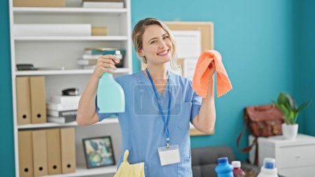 Téléchargez les photos : Jeune femme blonde nettoyant professionnel tenant chiffon et pulvérisateur au bureau - en image libre de droit