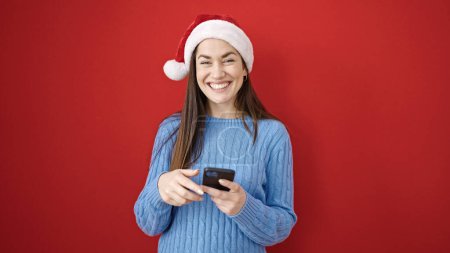 Foto de Mujer caucásica joven que usa sombrero de Navidad con teléfono inteligente sobre fondo rojo aislado - Imagen libre de derechos