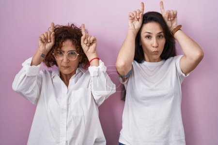 Photo for Hispanic mother and daughter wearing casual white t shirt over pink background doing funny gesture with finger over head as bull horns - Royalty Free Image