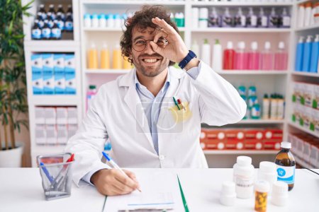 Téléchargez les photos : Jeune homme hispanique travaillant à la pharmacie faisant un geste correct avec la main souriante, les yeux regardant à travers les doigts avec un visage heureux. - en image libre de droit