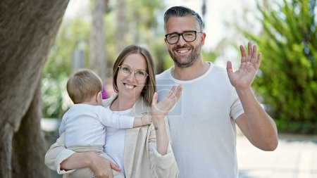 Téléchargez les photos : Famille de mère, père et bébé souriant ensemble regardant une caméra agitant au parc - en image libre de droit