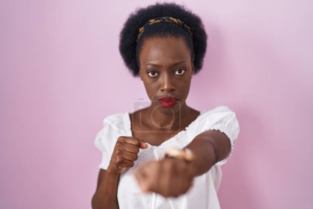 Téléchargez les photos : Femme africaine avec les cheveux bouclés debout sur fond rose poing poing pour se battre, agressif et en colère attaque, menace et violence - en image libre de droit