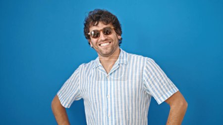 Téléchargez les photos : Jeune homme hispanique souriant confiant portant des lunettes de soleil sur fond bleu isolé - en image libre de droit