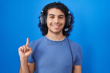 Téléchargez les photos : Homme hispanique aux cheveux bouclés écoutant de la musique à l'aide d'écouteurs montrant et pointant vers le haut avec le doigt numéro un tout en souriant confiant et heureux. - en image libre de droit