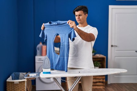 Photo for Hispanic man ironing holding burned iron shirt at laundry room skeptic and nervous, frowning upset because of problem. negative person. - Royalty Free Image