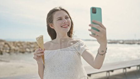 Photo for Young blonde woman tourist holding ice cream make selfie by smartphone at seaside - Royalty Free Image