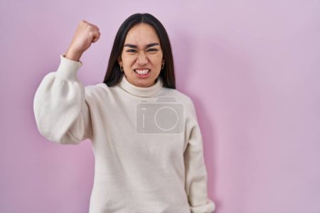 Téléchargez les photos : Jeune femme sud-asiatique debout sur fond rose en colère et fou levant le poing frustré et furieux tout en criant de colère. rage et concept agressif. - en image libre de droit
