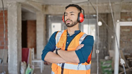 Photo for Young hispanic man builder with crossed arms wearing noise cancelling earmuffs at construction site - Royalty Free Image