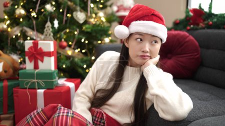 Photo for Young chinese woman sitting on floor by christmas tree with serious expression at home - Royalty Free Image