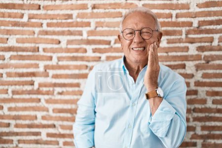Téléchargez les photos : Homme âgé avec les cheveux gris debout sur le mur de briques toucher la bouche avec la main avec une expression douloureuse en raison de maux de dents ou de maladies dentaires sur les dents. dentiste - en image libre de droit