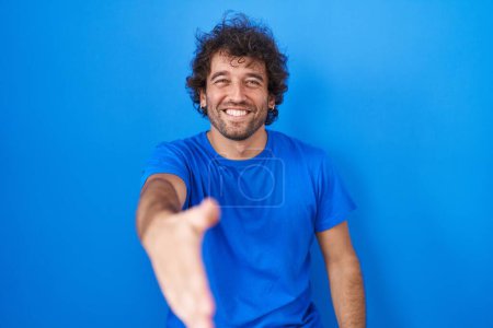 Foto de Joven hispano de pie sobre fondo azul sonriente amistoso ofreciendo apretón de manos como saludo y bienvenida. negocios exitosos. - Imagen libre de derechos