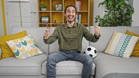 Téléchargez les photos : Jeune homme hispanique soutenant l'équipe de football assis sur le canapé à la maison - en image libre de droit