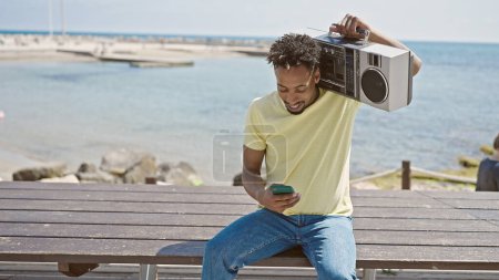 Téléchargez les photos : Africain américain tenant boombox en utilisant un smartphone au bord de la mer - en image libre de droit