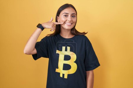 Téléchargez les photos : Jeune femme hispanique portant Bitcoin t-shirt souriant gai montrant et pointant avec les doigts dents et la bouche. concept de santé dentaire. - en image libre de droit