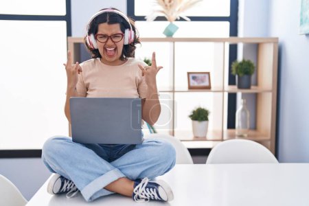 Photo for Young hispanic woman using laptop sitting on the table wearing headphones shouting with crazy expression doing rock symbol with hands up. music star. heavy concept. - Royalty Free Image