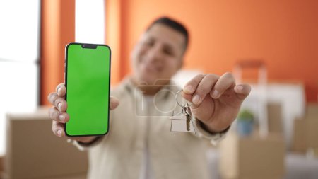 Photo for Young hispanic man showing screen smartphone holding keys at new home - Royalty Free Image