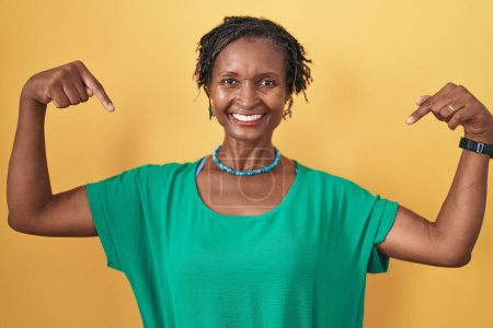 Foto de Mujer africana con rastas de pie sobre fondo amarillo mirando confiado con sonrisa en la cara, señalándose con los dedos orgullosos y felices. - Imagen libre de derechos