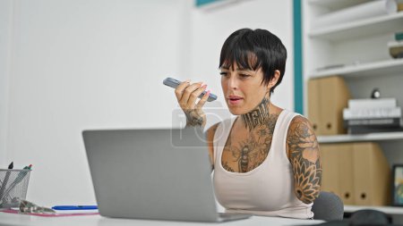 Téléchargez les photos : Femme hispanique avec un travailleur d'affaires amputé bras en utilisant un ordinateur portable envoyer un message vocal par smartphone au bureau - en image libre de droit