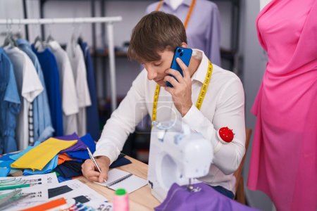 Téléchargez les photos : Jeune homme caucasien tailleur parler sur smartphone écriture sur ordinateur portable au magasin de tailleur - en image libre de droit