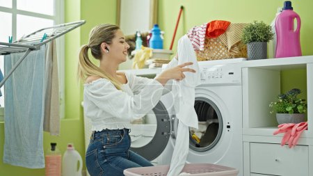 Photo for Young blonde woman listening to music washing clothes dancing at laundry room - Royalty Free Image