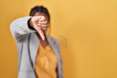 Téléchargez les photos : Femme afro-américaine avec des tresses debout sur fond jaune regardant malheureux et en colère montrant rejet et négatif avec les pouces vers le bas geste. mauvaise expression. - en image libre de droit