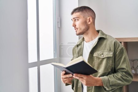 Photo for Young hispanic man reading book standing at home - Royalty Free Image