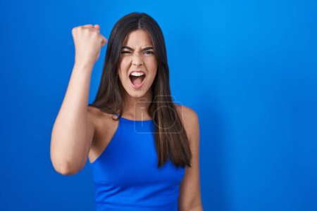 Téléchargez les photos : Femme hispanique debout sur fond bleu en colère et fou poing levant frustré et furieux tout en criant avec colère. rage et concept agressif. - en image libre de droit