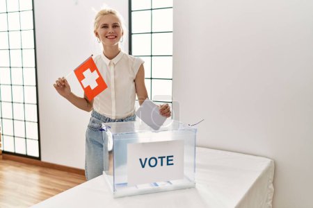 Foto de Joven rubia sosteniendo bandera suiza votando en colegio electoral - Imagen libre de derechos